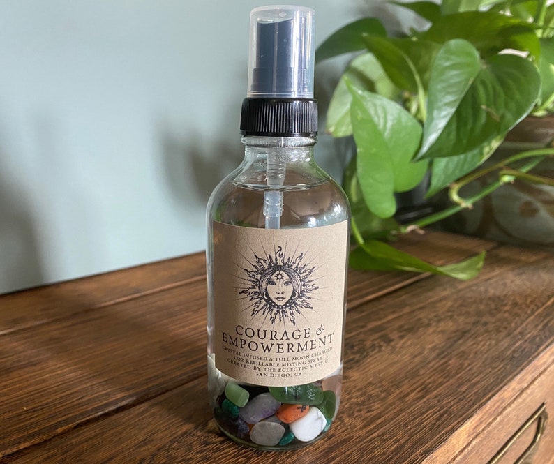 Puede incluir: Una botella de vidrio transparente con un pulverizador negro llena de un l&iacute;quido transparente y cristales. La botella tiene una etiqueta marr&oacute;n con el texto "COURAGE & EMPOWERMENT" y un sol con un dise&ntilde;o de cara.