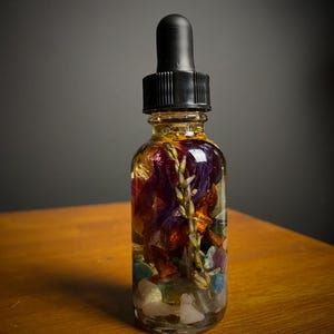 May include: A clear glass bottle with a black dropper top filled with a golden liquid and colorful dried flowers and crystals.