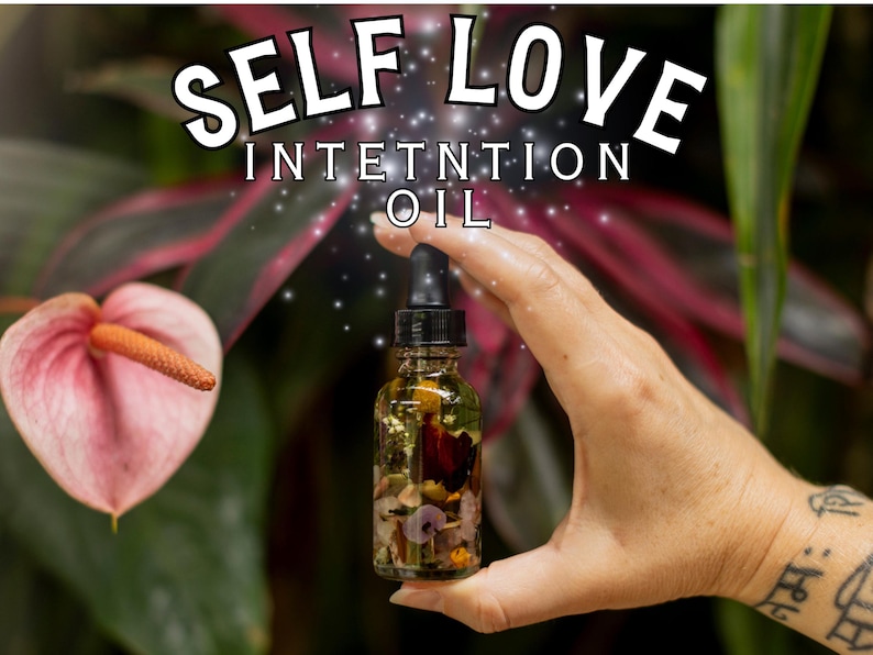 May include: A clear glass bottle filled with oil and botanicals, held in a hand. The bottle has a black dropper and is labeled "SELF LOVE INTENTION OIL". A pink flower and green foliage are in the background.