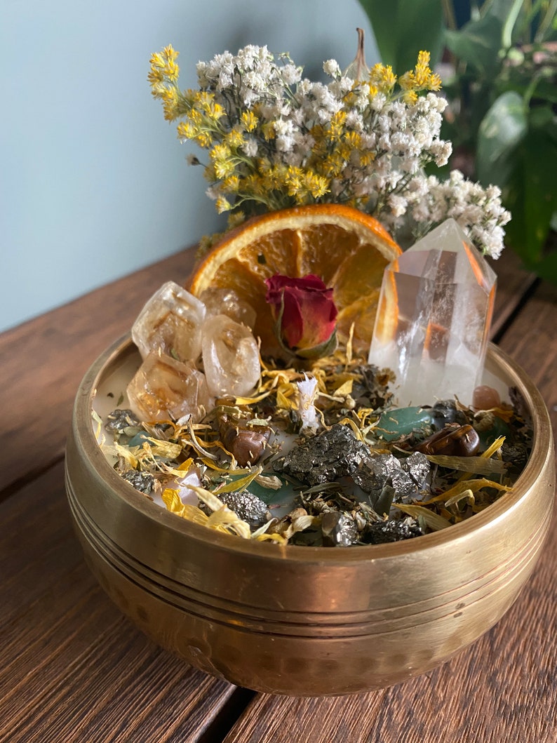 May include: A gold bowl filled with a white candle, dried flowers, orange slices, and various crystals. The bowl is sitting on a wooden surface.