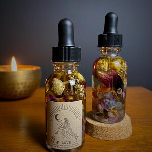 May include: Two small glass bottles filled with dried flowers and crystals. The bottles have black dropper tops and are labeled "SELF LOVE OIL" with a moon and star design. The bottles are sitting on a wooden surface.