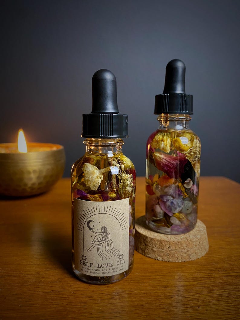 May include: Two small glass bottles filled with dried flowers and crystals. The bottles have black dropper tops and are labeled "SELF LOVE OIL" with a moon and star design. The bottles are sitting on a wooden surface.