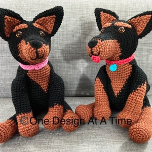 Puede incluir: Dos juguetes de peluche de perro tejidos a crochet, uno negro y marrón con un collar rosa y el otro negro y marrón con un collar rojo. Los perros están sentados en un sofá gris. El texto "One Design At A Time" es visible en la parte inferior central de la imagen.