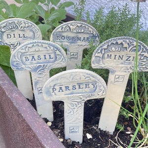 Könnte beinhalten: Fünf cremeweiße Keramik-Pflanzenstecker mit geprägten Blumenmustern und bläulichen Details. Jeder Stecker hat ein rechteckiges Etikettenfeld mit der Aufschrift DILL, ROSEMARY, BASIL, MINZE oder PARSLEY sowie ein kleines AJ-Logo am Stiel.
