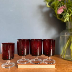 May include: Four ruby red wine glasses with clear stems and bases are arranged on a wooden surface. The glasses are in front of a vase of flowers. The glasses are approximately 10 cm tall.