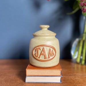 May include: A ceramic jar with a light brown glaze and the word "JAM" in brown lettering on the front. The jar is sitting on a stack of books.