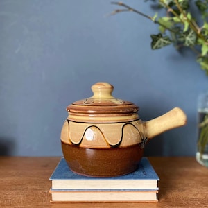 May include: A ceramic soup bowl with a lid and handle, in shades of brown and tan, with dark brown drip-like accents. The bowl is sitting on top of two blue books. The background is a blue wall and a wooden table.