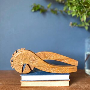 May include: A light brown, intricately carved wooden nutcracker, featuring floral and geometric patterns. It is placed on a stack of books. The background includes a blue wall and a vase with green foliage.