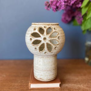 May include: A beige ceramic candle holder with a floral design cut-out. The candle holder has a round top and a cylindrical base. It sits on a small book. Purple flowers are in the background.