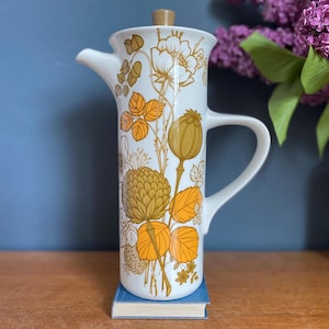 May include: A tall, white ceramic coffee pot with a floral design in gold and orange. The pot has a spout, handle, and a lid. The floral pattern includes flowers, leaves, and seed pods. The pot is sitting on a small blue book.