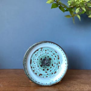 May include: A ceramic bowl with a light blue exterior and a turquoise interior with brown speckles. The bowl has a circular design and sits on a wooden surface. The background features a blue wall and a glass vase with greenery.