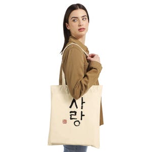 Könnte beinhalten: Cremefarbene Tragetasche mit schwarzen koreanischen Schriftzeichen und einem roten quadratischen Stempel. Die Tasche wird über der Schulter getragen. Die Person trägt eine braune Jacke und blaue Jeans.