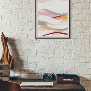 May include: A framed abstract painting with a white background and colorful brushstrokes in shades of pink, yellow, gray, and red. The painting is hanging on a white brick wall above a wooden desk with a laptop, camera, and a wooden tray.