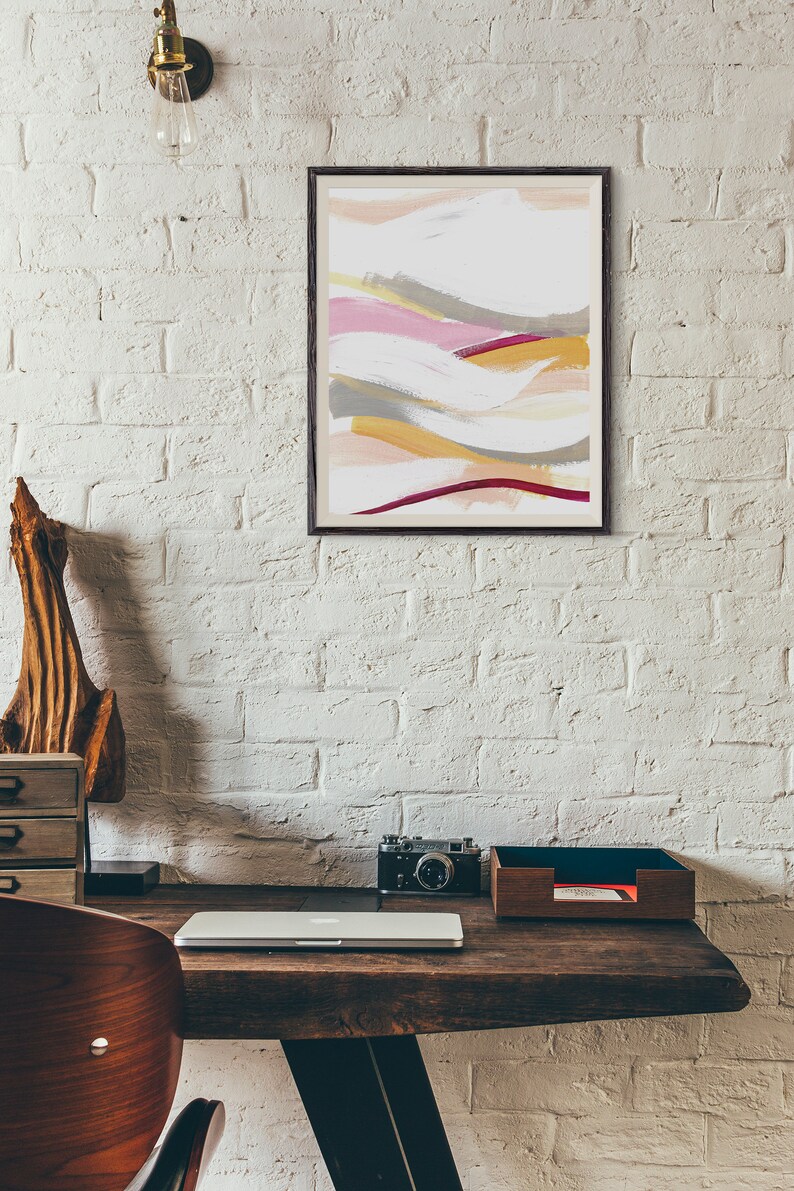 May include: A framed abstract painting with a white background and colorful brushstrokes in shades of pink, yellow, gray, and red. The painting is hanging on a white brick wall above a wooden desk with a laptop, camera, and a wooden tray.