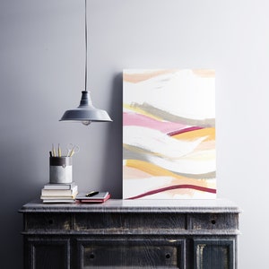 May include: Abstract painting with a white background and colorful brushstrokes in shades of pink, yellow, and gray. The painting is hanging on a wall above a dark wooden dresser.