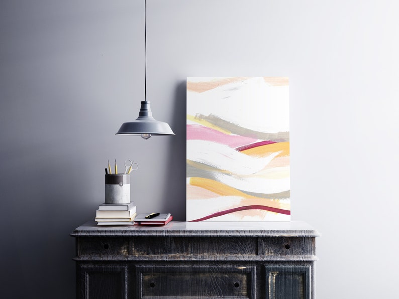 May include: Abstract painting with a white background and colorful brushstrokes in shades of pink, yellow, and gray. The painting is hanging on a wall above a dark wooden dresser.