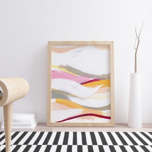May include: Abstract painting in a wooden frame with pink, yellow, orange, and gray brushstrokes on a white background. The painting is hanging on a white wall above a wooden table and a white vase with a dried branch.
