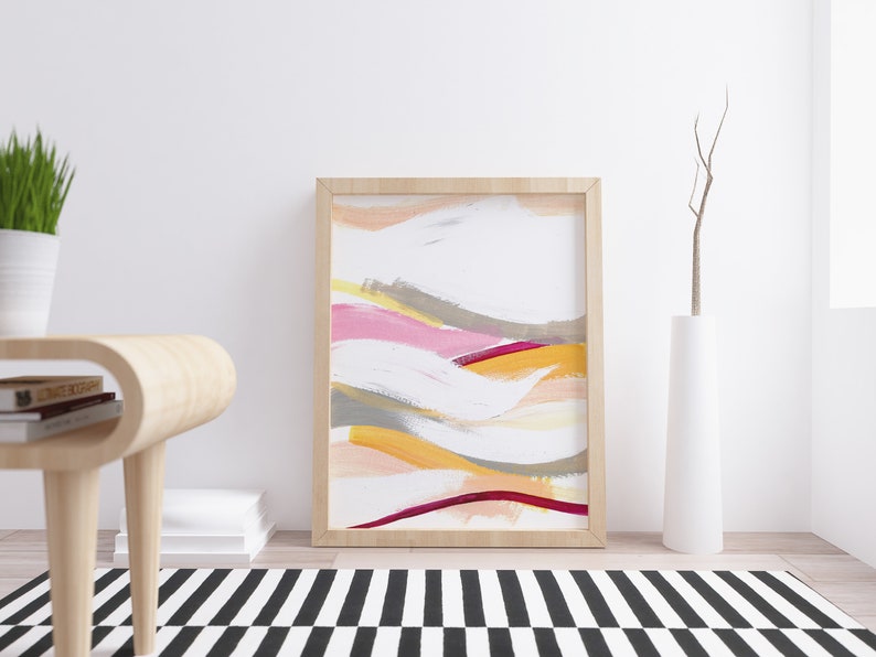 May include: Abstract painting in a wooden frame with pink, yellow, orange, and gray brushstrokes on a white background. The painting is hanging on a white wall above a wooden table and a white vase with a dried branch.