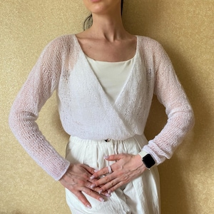 Pu&ograve; includere: Un maglione a portafoglio bianco a maniche lunghe con una trama a maglia aperta e trasparente. Il capo presenta un profondo scollo a V che si sovrappone sul davanti, maniche lunghe e una silhouette corta, indossato sopra una canotta bianca.