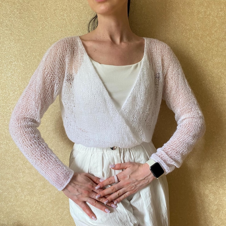 Pu&ograve; includere: Un maglione a portafoglio bianco a maniche lunghe con una trama a maglia aperta e trasparente. Il capo presenta un profondo scollo a V che si sovrappone sul davanti, maniche lunghe e una silhouette corta, indossato sopra una canotta bianca.
