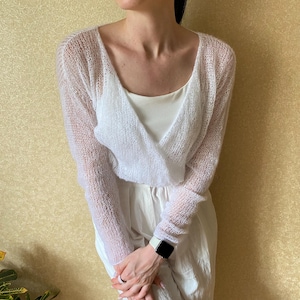 Pu&ograve; includere: Un maglione a portafoglio bianco a maniche lunghe realizzato in materiale trasparente a maglia larga. Il capo presenta un profondo scollo a V e una silhouette corta, indossato sopra una canotta bianca a coste e pantaloni bianchi.