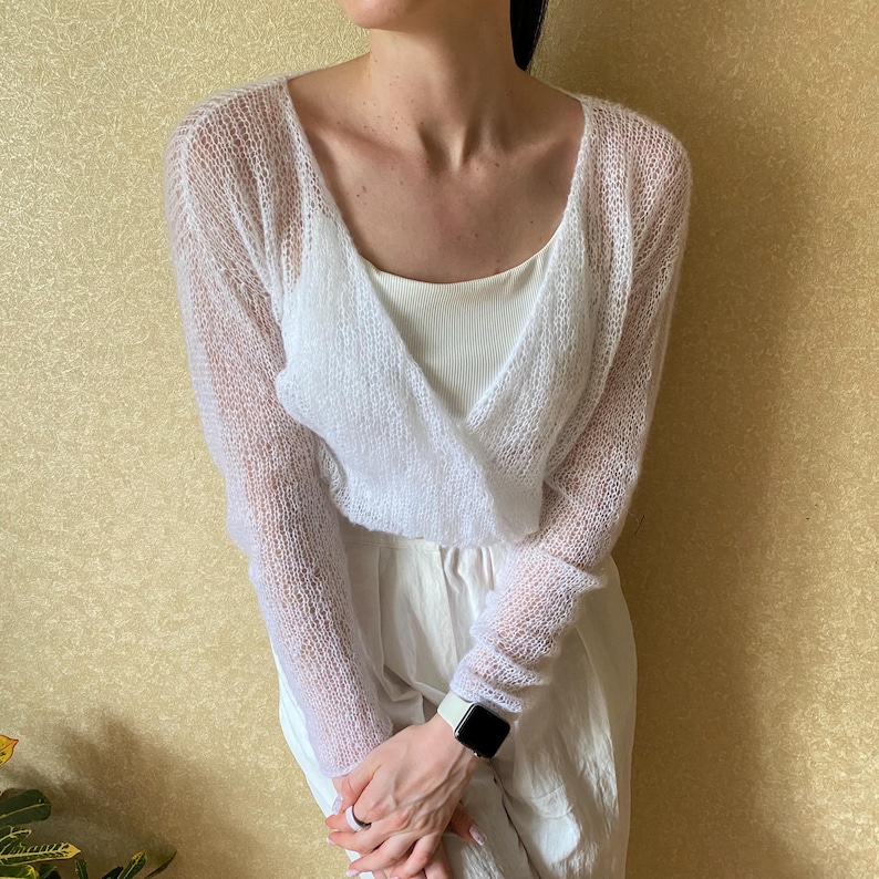 Pu&ograve; includere: Un maglione a portafoglio bianco a maniche lunghe realizzato in materiale trasparente a maglia larga. Il capo presenta un profondo scollo a V e una silhouette corta, indossato sopra una canotta bianca a coste e pantaloni bianchi.