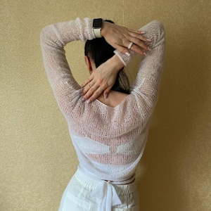 Pu&ograve; includere: Un maglione a maniche lunghe in maglia color bianco sporco, trasparente e con una trama a maglia larga. Il capo presenta un'ampia scollatura e una chiusura a fiocco in vita, indossato sopra un indumento intimo bianco con pantaloni bianchi.