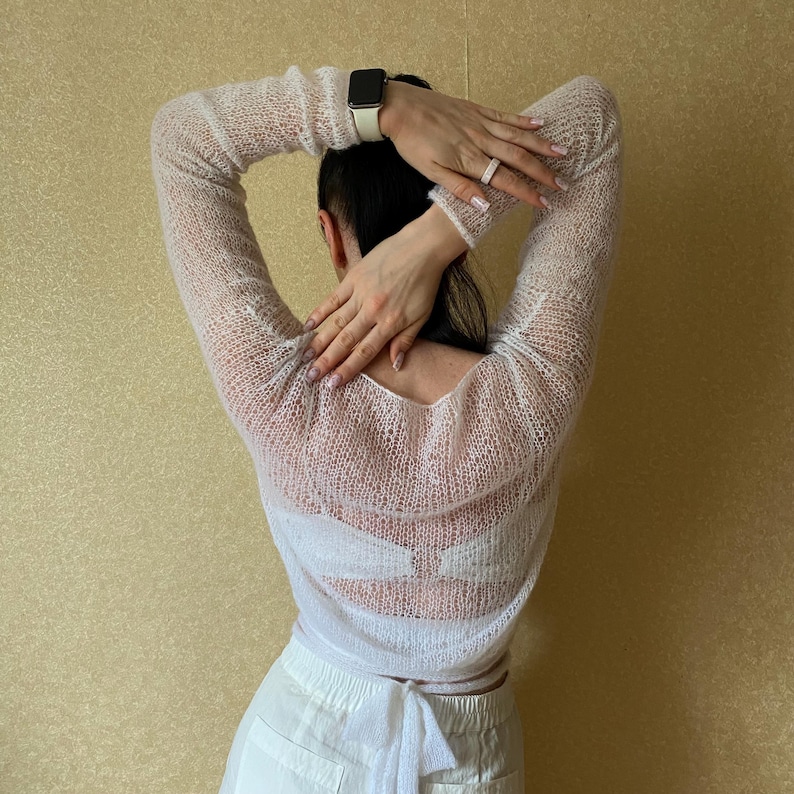 Pu&ograve; includere: Un maglione a maniche lunghe in maglia color bianco sporco, trasparente e con una trama a maglia larga. Il capo presenta un'ampia scollatura e una chiusura a fiocco in vita, indossato sopra un indumento intimo bianco con pantaloni bianchi.