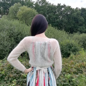 Pu&ograve; includere: Un maglione in maglia trasparente color bianco sporco, a maniche lunghe e con chiusura a fiocco in vita. Il maglione &egrave; indossato sopra un abito senza maniche a righe verticali nei toni del rosso, blu, nero e bianco, con un fiocco visibile sul retro.