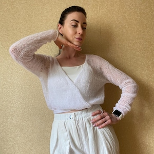 Puede incluir: Un suéter cruzado blanco de manga larga con una textura de punto abierto y transparente. El suéter se lleva sobre una camiseta blanca lisa, combinado con pantalones blancos de talle alto con cierre de botón y diseño plisado frontal.