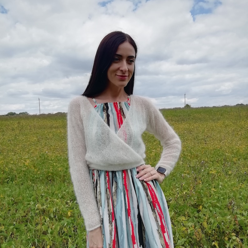 Pu&ograve; includere: Un cardigan a portafoglio corto a maniche lunghe di colore bianco, realizzato in un materiale a maglia soffice e trasparente. Il capo presenta uno scollo a V ed &egrave; indossato sopra un abito senza maniche con righe verticali rosse, nere, azzurre e bianche.