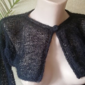 Può includere: Un bolero corto nero caratterizzato da una trama trasparente a maglia aperta, maniche lunghe e una chiusura con un unico bottone circolare sullo scollo. Il capo è realizzato in filato leggero e soffice con una lavorazione a maglia larga.