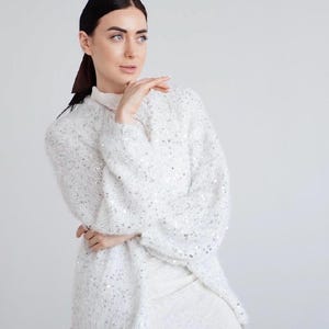Può includere: Un maglione bianco a maniche lunghe in maglia con collo alto e paillettes argentate sparse su tutto il tessuto. Il maglione presenta una trama testurizzata e ampia, indossato sopra un abito bianco con un sottile motivo geometrico ripetuto.
