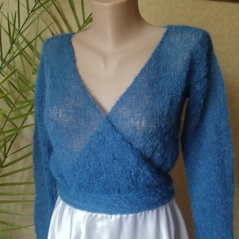 Pu&ograve; includere: Un maglione a portafoglio blu a maniche lunghe con un profondo scollo a V e una consistenza lavorata a maglia soffice e semi-trasparente. Il capo &egrave; aderente in vita con una fascia larga ed &egrave; esposto su un manichino, mostrando la trama aperta.