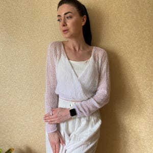Pu&ograve; includere: Un cardigan a portafoglio bianco a maniche lunghe realizzato in un tessuto a maglia fine e trasparente. Il capo presenta uno scollo a V e una silhouette corta, indossato sopra una canotta bianca tinta unita e abbinato a pantaloni bianchi.