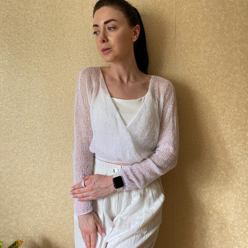 Pu&ograve; includere: Un cardigan a portafoglio bianco a maniche lunghe realizzato in un tessuto a maglia fine e trasparente. Il capo presenta uno scollo a V e una silhouette corta, indossato sopra una canotta bianca tinta unita e abbinato a pantaloni bianchi.