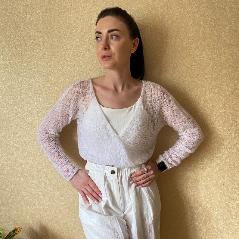 Pu&ograve; includere: Un coprispalle bianco a maniche lunghe in stile avvolgente, realizzato in un tessuto a maglia leggero e trasparente. Il capo presenta un profondo scollo a V, maniche aderenti e una lunghezza corta in vita, indossato sopra un top bianco.