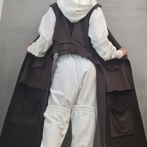 May include: A white hooded sweatshirt with gathered sleeves, white sweatpants featuring gathered knee sections, and a long, sleeveless dark brown vest with large patch pockets. The outfit is paired with black sneakers with thick white soles.
