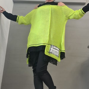May include: A neon yellow loose-knit sweater with an off-the-shoulder neckline, layered over a black top and black lace hem. The sweater features a rectangular patch with the text "INFORMAT" and black leggings paired with black boots with silver chains.