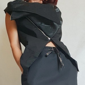 Pode incluir: Top preto sem mangas com um design assimétrico e um zíper diagonal. O top é combinado com calças pretas. O traje tem uma estética moderna e ousada.