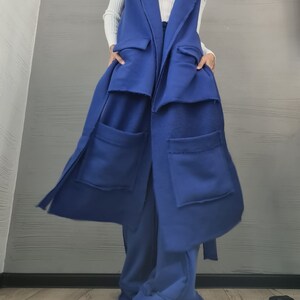 May include: A long, sleeveless royal blue vest featuring a wide lapel, two large patch pockets on the lower front, and two smaller flap pockets at the waist. The garment is constructed from a thick, fleece-like fabric with visible raw edges.