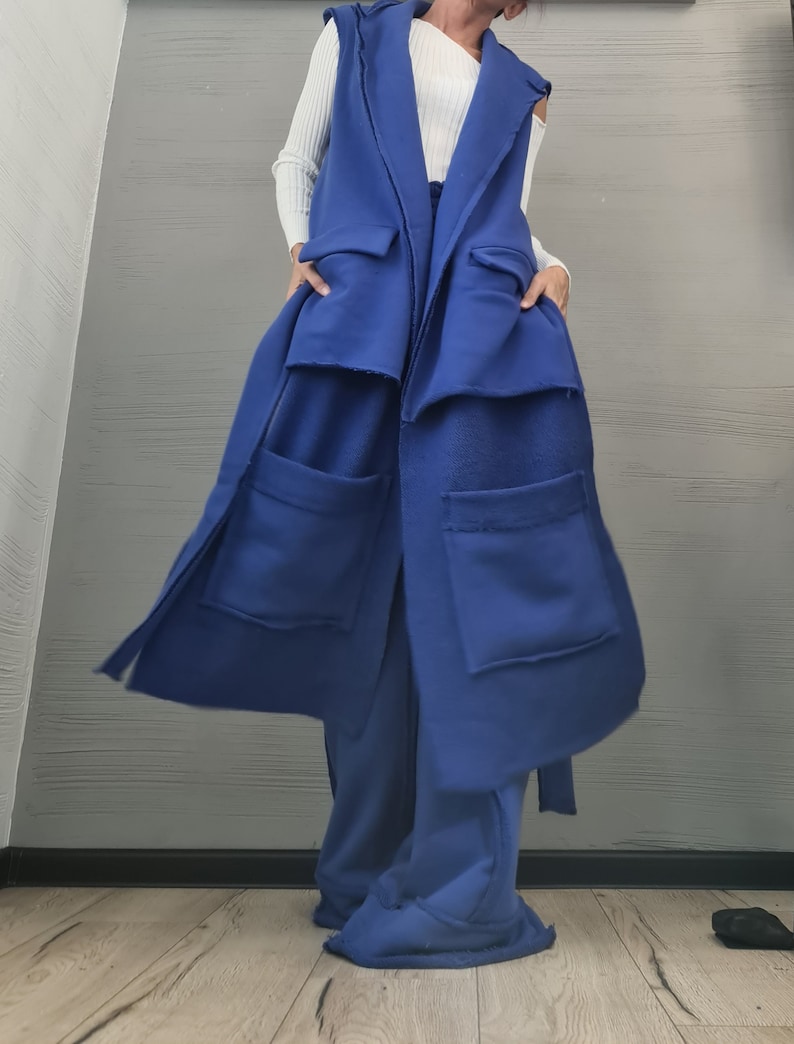 May include: A long, sleeveless royal blue vest featuring a wide lapel, two large patch pockets on the lower front, and two smaller flap pockets at the waist. The garment is constructed from a thick, fleece-like fabric with visible raw edges.
