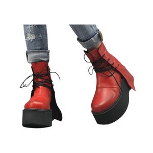 K&ouml;nnte beinhalten: Rote Plateau-Stiefel mit schwarzen Schn&uuml;rsenkeln und schwarzer Sohle. Die Stiefel haben ein einzigartiges Design mit einer Klappe an der Seite. Die Stiefel werden mit blauen Jeans getragen.