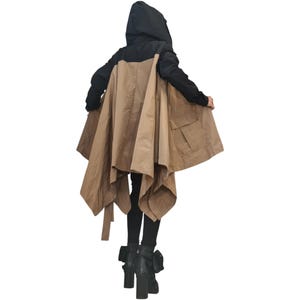 Op de afbeelding: Een jas met capuchon met een zwart bovengedeelte en een beige ondergedeelte met een uitlopende, asymmetrische zoom. Het kledingstuk heeft lange zwarte mouwen, een capuchon en een grote zak aan de zijkant. Gecombineerd met een zwarte broek en zwarte laarzen.