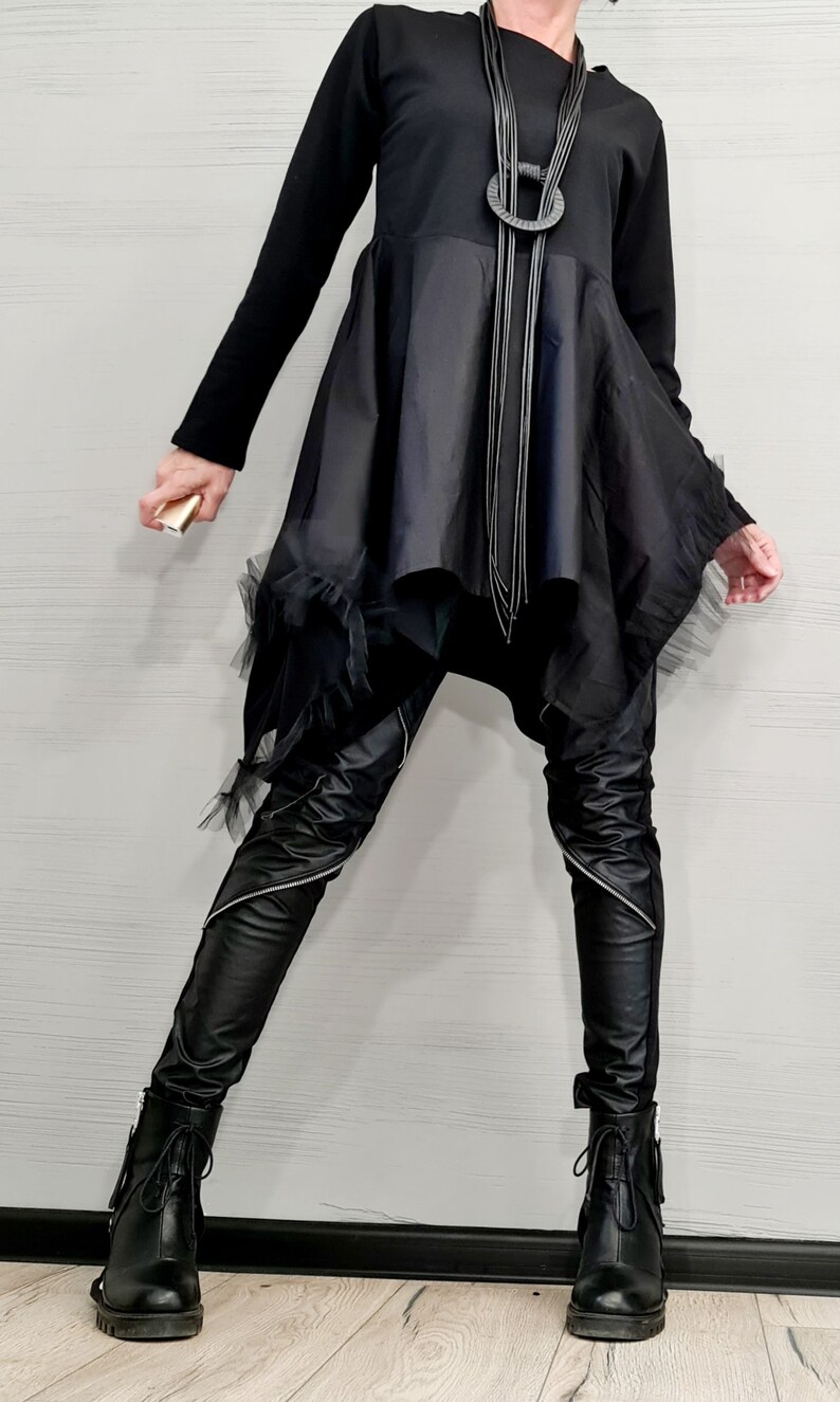 May include: Black asymmetrical tunic dress with a long-sleeved top and a flowing skirt. The outfit includes black leather-look pants with zippers, black boots, and a layered necklace. A small gold-colored object is held.