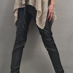 May include: A sleeveless beige knit turtleneck top with a loose, asymmetrical hem, paired with black faux leather pants featuring diagonal silver zippers on the legs. A long black cord necklace with a large circular pendant and black buckled boots.