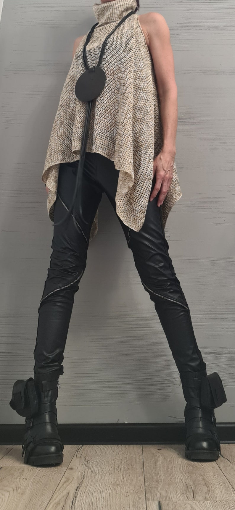 May include: A sleeveless beige knit turtleneck top with a loose, asymmetrical hem, paired with black faux leather pants featuring diagonal silver zippers on the legs. A long black cord necklace with a large circular pendant and black buckled boots.