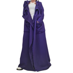 May include: A long, deep purple hooded coat featuring a draped open front, long sleeves, and two prominent circular cut-out openings on the upper chest area. The coat is worn over a simple white sleeveless top and reaches down to the floor.