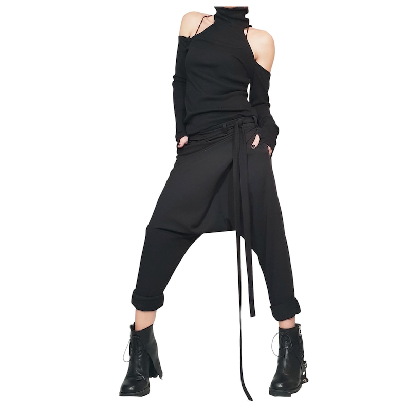 May include: Black ensemble featuring a long-sleeved top with shoulder cutouts and a high neckline. The outfit includes draped, loose-fitting pants with a wrap-style design and a long tie. The model is wearing black ankle boots.