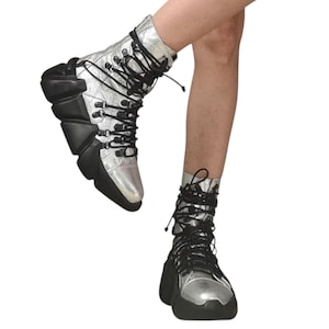 May include: Silver and black high-top boots with a unique, geometric design. The boots feature a metallic silver upper with black accents on the sole and heel. Black laces weave up the front, adding a contrasting detail to the futuristic footwear.